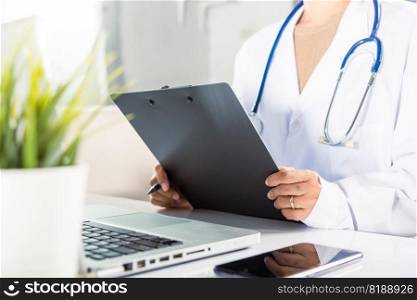 Closeup of doctor or nurse woman in uniform with stethoscope writing information of patient prescription in paperwork on clipboard and typing laptop computer for history record medical document report