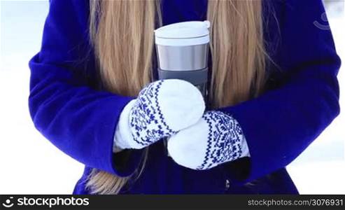 Closeup of female hands in knitted mittens with hot drink in metal thermal mug outdoors in winter. Woman&acute;s hands offering hot coffee in thermocup in cold wintertime.
