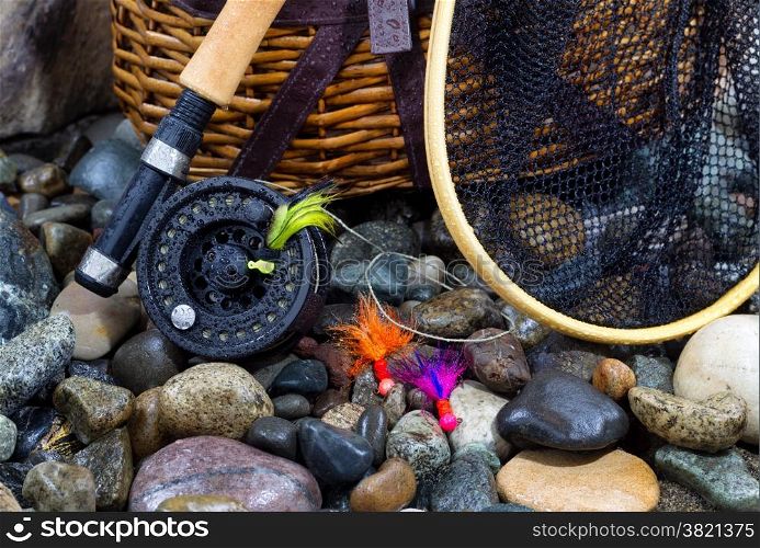 Closeup of fishing fly reel, landing net, creel and assorted flies on wet river bed stones&#xA;