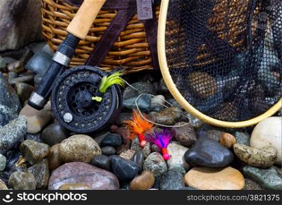 Closeup of fishing fly reel, landing net, creel and assorted flies on wet river bed stones&#xA;