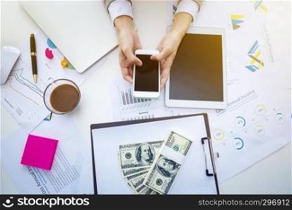 Closeup of hand holding mobile on desk with paperwork, money and office accessories. Business and technology concept.