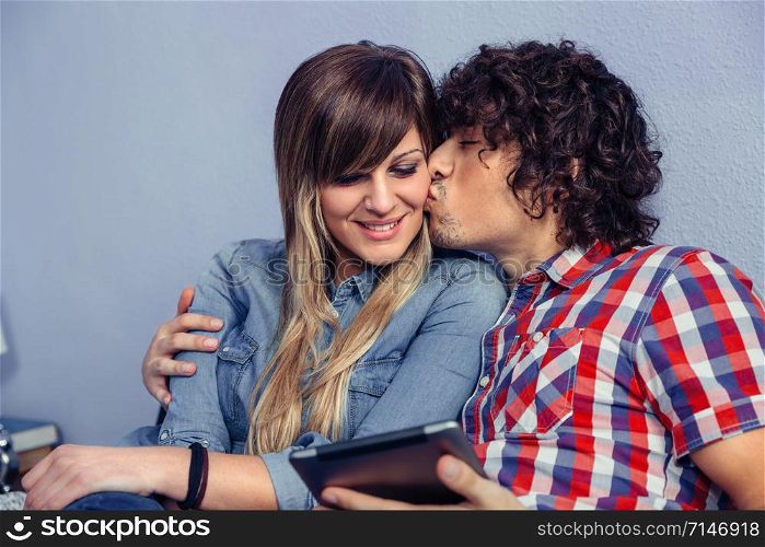 Closeup of handsome man kissing to young beautiful woman and holding electronic tablet in his hand. Love and leisure time concept.. Man kissing to young woman and holding tablet