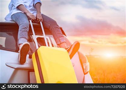 Closeup of happy Asian woman on top of car with luggage bag. Girl sitting on roof and looking sunset before evening. People lifestyle in long vacation trip concept. Nature and transportation vehicle