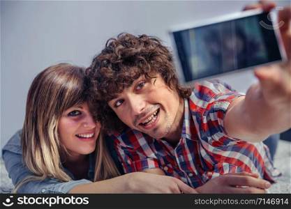 Closeup of happy young couple in love taking a selfie with smartphone lying over a bed. Leisure time at home concept.. Couple in love taking selfie with smartphone on bed