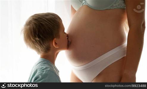 Closeup of little boy kissing his pregnatn mom in big belly standing at window in bedroom. Concept of family happiness and baby anticipation.. Closeup of little boy kissing his pregnatn mom in big belly standing at window in bedroom. Concept of family happiness and baby anticipation