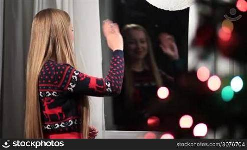 Closeup of lovely cheerful woman looking out of the window and waving hello to her boyfriend on Christmas eve. Charming blonde girl standing in evening and welcoming her lover during celebration of winter holidays together at home.