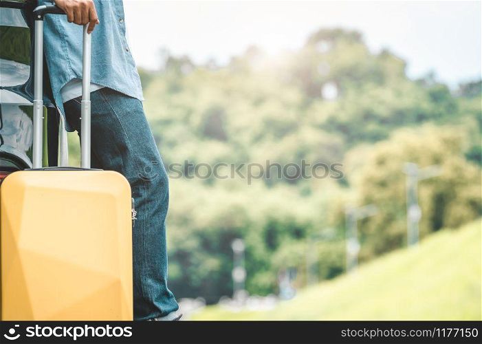Closeup of man legs resting during travel on road trip with yellow suitcase on summer nature background. Transportation and people lifestyles concept. Tourist standing outdoors on vacation tour