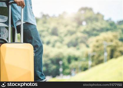 Closeup of man legs resting during travel on road trip with yellow suitcase on summer nature background. Transportation and people lifestyles concept. Tourist standing outdoors on vacation tour