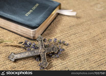 Closeup of metalic Christian cross on lavanda flowers next to Holy Bible on textile background.