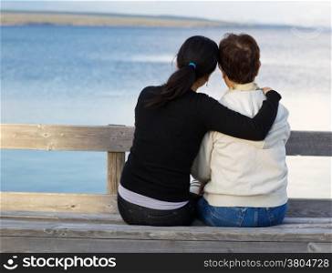 Closeup of mother with mature daughter holding her while sitting on wooden bench looking outward at the lake