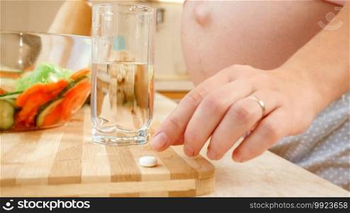 Closeup of pregnant woman taking vitamin pill and drinking glass of clear water before breakfast on kitchen. Concept of healthy lifestyle, nutrition and hydration during pregnancy.. Closeup of pregnant woman taking vitamin pill and drinking glass of clear water before breakfast on kitchen. Concept of healthy lifestyle, nutrition and hydration during pregnancy