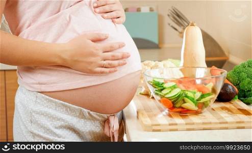 Closeup of pregnant woman with big belly standing on kitchen and touching big abdomen. Concept of healthy lifestyle and nutrition during pregnancy.. Closeup of pregnant woman with big belly standing on kitchen and touching big abdomen. Concept of healthy lifestyle and nutrition during pregnancy