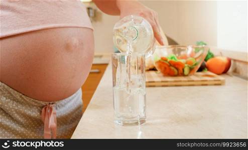 Closeup of pregnant young woman pouring water in glass and drinking it on kitchen at morning. Concept of healthy lifestyle, nutrition and hydration during pregnancy.. Closeup of pregnant young woman pouring water in glass and drinking it on kitchen at morning. Concept of healthy lifestyle, nutrition and hydration during pregnancy