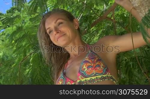 Closeup of pretty blonde woman wearing colourful swimwear sitting on a branch on a beautiful summer day over green leaves background - video in slow motion