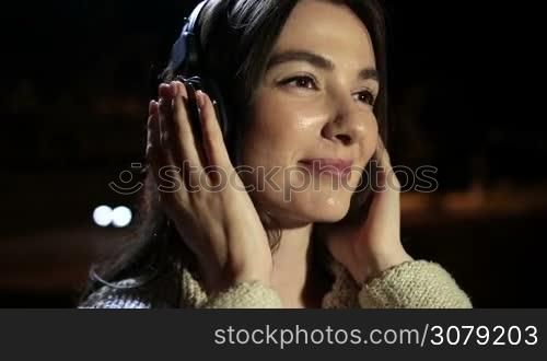 Closeup of romantic girl with headphones listening to music as she enjoys leisure on a warm summer night outdoors. Millennial brunette woman in earphones relaxing in night city, swinging along with the beat of favorite song on streetlights bokeh