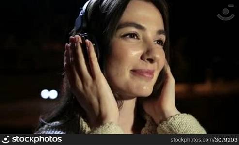 Closeup of romantic girl with headphones listening to music as she enjoys leisure on a warm summer night outdoors. Millennial brunette woman in earphones relaxing in night city, swinging along with the beat of favorite song on streetlights bokeh