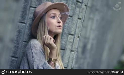 Closeup of sad lonely woman smoking cigarette and thinking. Upset woman smoking under the stress and leaning on the loft brick wall.