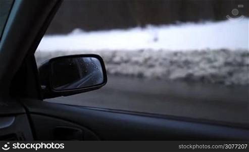 Closeup of side rearview mirror of car while driving on highway on a winter day. Drops of melted snow on car side view mirror during driving in wintertime and passing traffic on background.