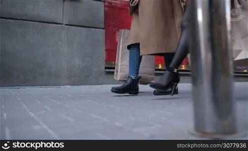 Closeup of slender females legs in high heels and mini dress walking along the street against store showcase background. Slow motion. Side view. Beautiful fashionable women carrying shopping bags going down cobblestone sidewalk after day shopping.