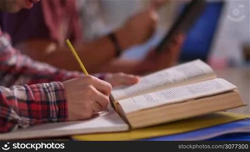 Closeup of student&acute;s male hands with book writing in notebook with pencil. Young male student studying together with friends at home, reading textbook and making notes in exercise book with pencil. Blurred students working on touchpad on hebakcground