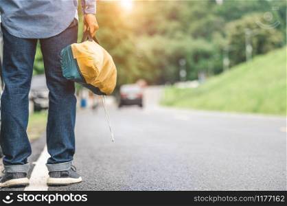 Closeup of tourist legs walking along road with bag during travel in countryside. People lifestyles and vacation concept. Man holding and backpacking for long holiday trip with mountain background