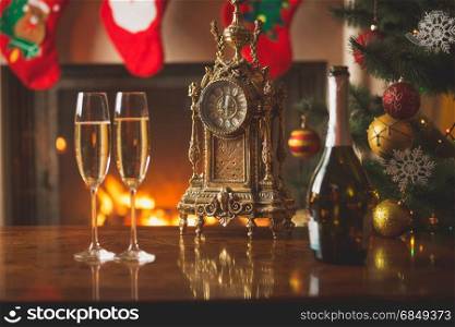 Closeup of two glasses of champagne on table next to old clock showing midnight at living room decorated for Christmas