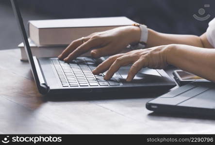 Closeup of woman hands typing on laptop keyboard with books and various devices on wooden table in home office room