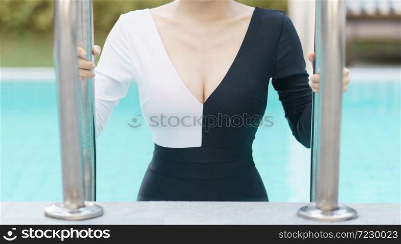 Closeup of Woman in black and white swimsuit posing at pool outdoors, holding stairs of swimming pool.