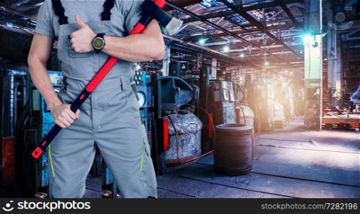 Closeup of worker man with ax dressed in overalls on Industrial zone background, manufacturing facility. Concept of Safe labor in the enterprise.. Industrial zone background