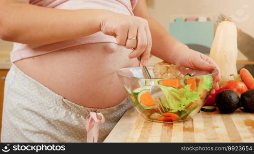 Closeup of yougn pregnant woman with big belly eating fresh vegetable salad while standing on kitchen. Concept of healthy lifestyle and nutrition during pregnancy.. Closeup of yougn pregnant woman with big belly eating fresh vegetable salad while standing on kitchen. Concept of healthy lifestyle and nutrition during pregnancy