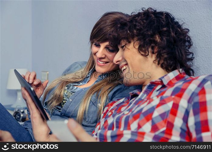 Closeup of young couple in love looking electronic tablet and laughing while resting over a bed. Leisure time at home concept.. Couple in love looking electronic tablet and laughing