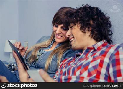 Closeup of young couple in love looking electronic tablet and laughing while resting over a bed. Leisure time at home concept.. Couple in love looking electronic tablet and laughing