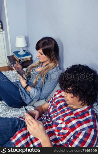 Closeup of young couple in love using electronic devices while resting over a bed. Leisure time at home concept.. Couple in love using electronic devices on bed