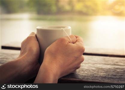 Closeup on a woman&rsquo;s hands holding a cup of tea by a lake in the afternoon