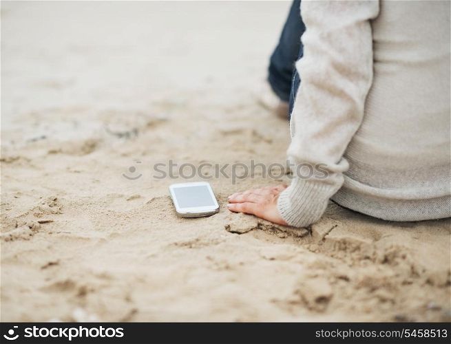 Closeup on cell phone near young woman in sweater sitting on beach