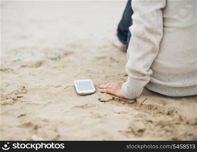 Closeup on cell phone near young woman in sweater sitting on beach
