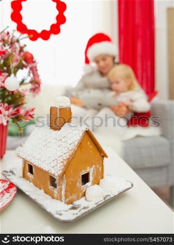 Closeup on Christmas Gingerbread house and mother and baby using tablet PC in background