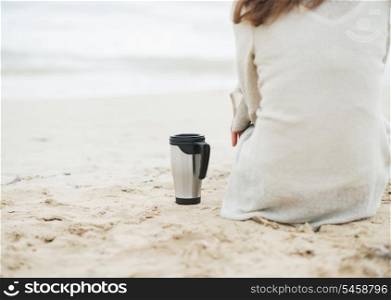 Closeup on cup of hot beverage near young woman in sweater sitting on lonely beach