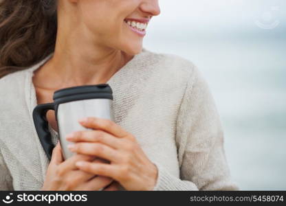 Closeup on happy young woman in sweater on beach with cup of hot beverage