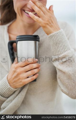 Closeup on happy young woman in sweater on beach with cup of hot beverage