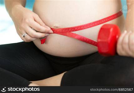 Closeup ot young pregnant woman doing fitness and measuring belly with measuring tape