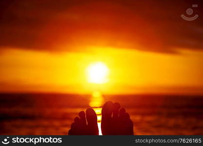 Closeup photo of a women&rsquo;s feet over background of a beautiful orange sunset over sea, pleasure and relaxation, summer vacation concept