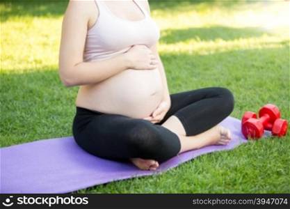 Closeup photo of active pregnant woman sitting on grass at park and touching big tummy
