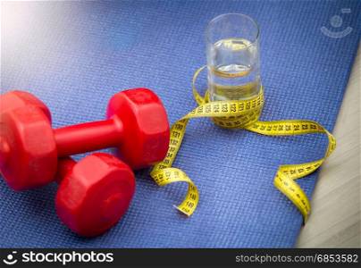 Closeup photo of dumbbells, glass of water and measuring tape on fitness mat next to woman doing exercises