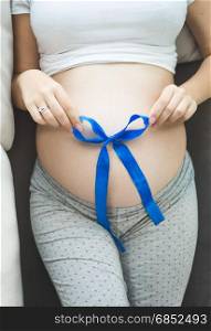 Closeup photo of pregnant woman lying on sofa and holding blue ribbon on belly