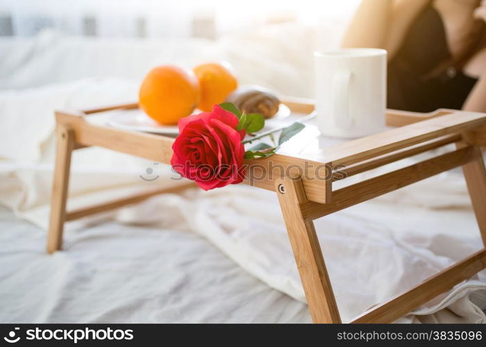 Closeup photo of tray with breakfast and red rose on bed at hotel room