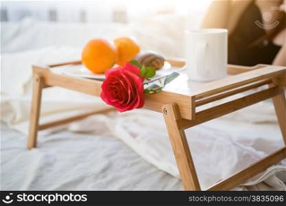 Closeup photo of tray with breakfast and red rose on bed at hotel room