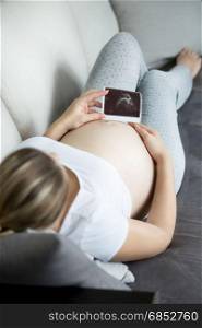 Closeup photo of young pregnant woman relaxing on sofa and looking at ultrasound baby scan