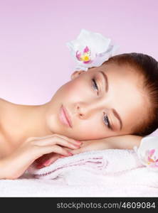 Closeup portrait of a beautiful woman, face with no makeup and perfect clear skin, female with a white orchid flower, lying down on massage table, enjoying a relaxing day at spa, pink background