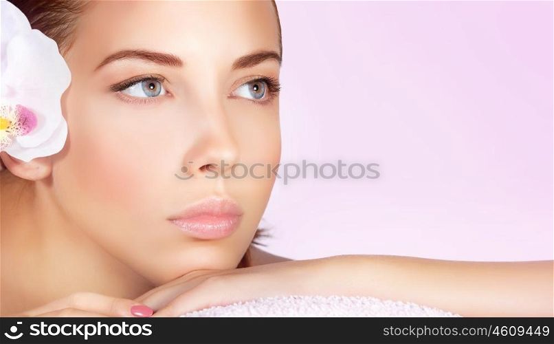 Closeup portrait of a beautiful woman, perfect face with no makeup makeup, female lying down on massage table white orchid flower in hair, enjoying day spa, shot over pink background with copy space